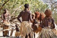 A group of people wearing traditional attire are gathered in an outdoor setting with a vibrant natural backdrop. The individuals are dressed in skirts made from natural materials and have unique hairstyles. One person is playing a large drum while others appear to be dancing or engaging in a ritual.