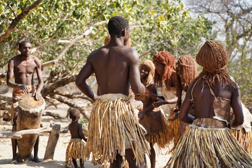 A group of people wearing traditional attire are gathered in an outdoor setting with a vibrant natural backdrop. The individuals are dressed in skirts made from natural materials and have unique hairstyles. One person is playing a large drum while others appear to be dancing or engaging in a ritual.