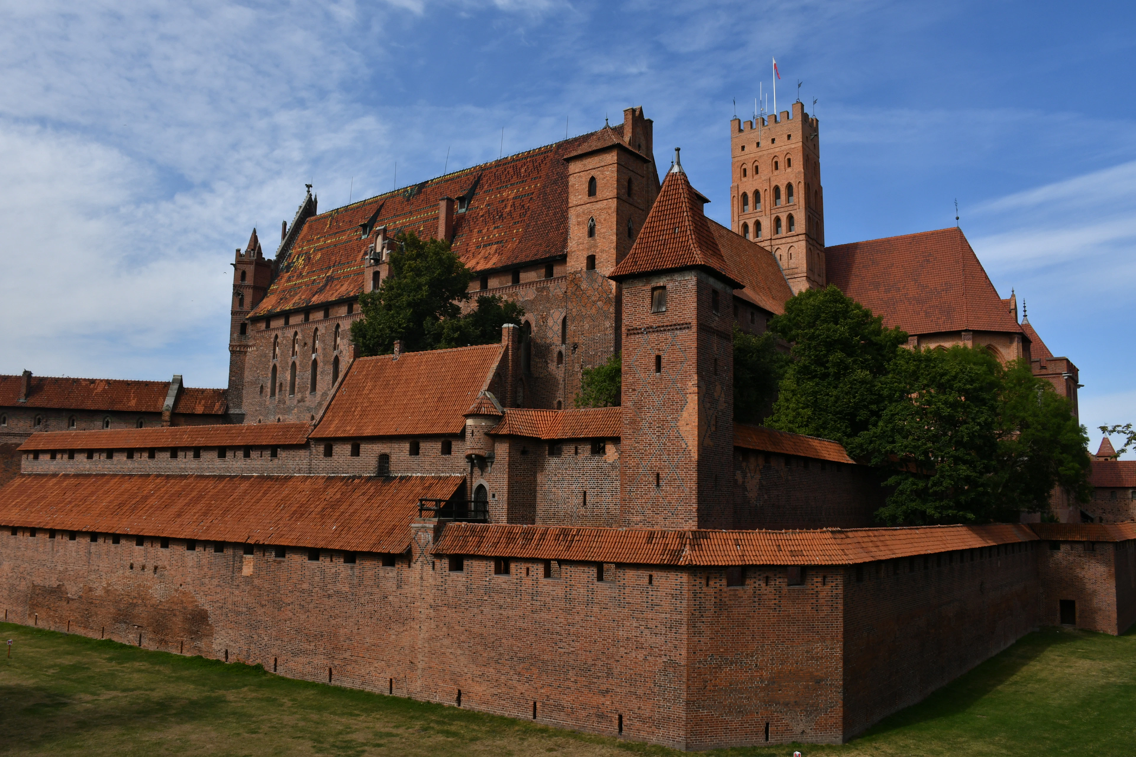Malbork Castle in full: tier on tier of red-tiled roofs and red brick rising behind the crenellated curtain wall and its corner tower, the Teutonic Knights' great fortress standing over a green sward under a deep blue summer sky.