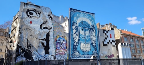 Several large murals cover the sides of buildings in an urban setting. One mural features a grayscale depiction of an exaggerated face with wide eyes, while another displays a colorful cat with a variety of vibrant hues. A blue-toned artwork presents a woman with a complex design, holding a book inscribed with 'The Future is Unwritten.' Another area features a pixelated pattern resembling a video game character.