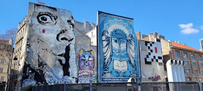 Several large murals cover the sides of buildings in an urban setting. One mural features a grayscale depiction of an exaggerated face with wide eyes, while another displays a colorful cat with a variety of vibrant hues. A blue-toned artwork presents a woman with a complex design, holding a book inscribed with 'The Future is Unwritten.' Another area features a pixelated pattern resembling a video game character.