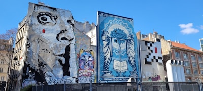 Several large murals cover the sides of buildings in an urban setting. One mural features a grayscale depiction of an exaggerated face with wide eyes, while another displays a colorful cat with a variety of vibrant hues. A blue-toned artwork presents a woman with a complex design, holding a book inscribed with 'The Future is Unwritten.' Another area features a pixelated pattern resembling a video game character.