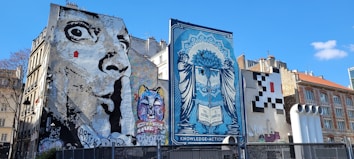 Several large murals cover the sides of buildings in an urban setting. One mural features a grayscale depiction of an exaggerated face with wide eyes, while another displays a colorful cat with a variety of vibrant hues. A blue-toned artwork presents a woman with a complex design, holding a book inscribed with 'The Future is Unwritten.' Another area features a pixelated pattern resembling a video game character.
