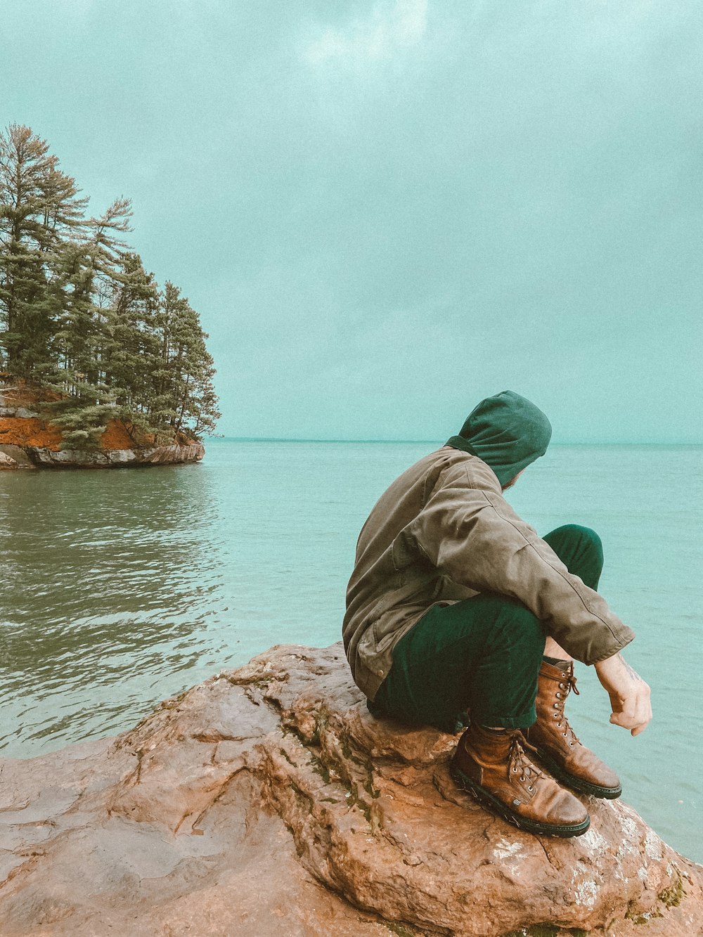 녹색 까마귀와 갈색 바지를 입은 남자가 수역 근처의 갈색 바위에 앉아 있습니다.