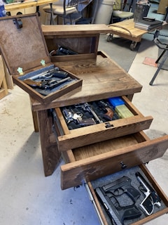 A wooden cabinet with multiple open drawers containing various firearms. The setting appears to be a workshop or garage with tools and equipment visible in the background. The cabinet features engraved designs.