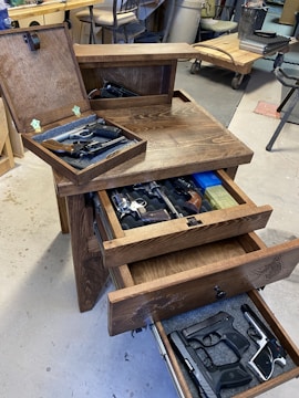A wooden cabinet with multiple open drawers containing various firearms. The setting appears to be a workshop or garage with tools and equipment visible in the background. The cabinet features engraved designs.