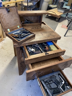A wooden cabinet with multiple open drawers containing various firearms. The setting appears to be a workshop or garage with tools and equipment visible in the background. The cabinet features engraved designs.