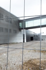 A metal fence is in the foreground with blurred view of a modern industrial or office building in the background. The structure features a metallic facade and a glass walkway connecting separate parts of the building. The area appears to be devoid of people.