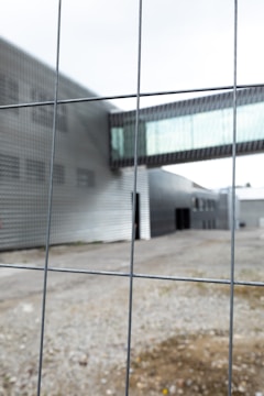 A metal fence is in the foreground with blurred view of a modern industrial or office building in the background. The structure features a metallic facade and a glass walkway connecting separate parts of the building. The area appears to be devoid of people.