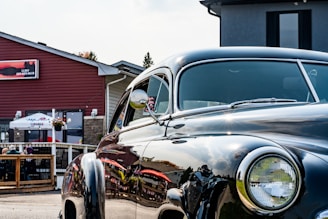 A polished vintage car is parked in front of a building with a patio where people are seated under a large white umbrella. The car's shiny surface reflects the surroundings, including the red-brown building and the blue sky. The building has a rustic design, and there are potted plants decorating the space.