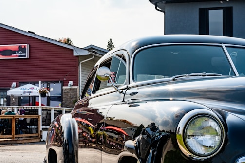 A polished vintage car is parked in front of a building with a patio where people are seated under a large white umbrella. The car's shiny surface reflects the surroundings, including the red-brown building and the blue sky. The building has a rustic design, and there are potted plants decorating the space.