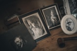 A collection of framed black and white photographs is displayed on a wooden surface. The frames contain portraits: a bearded old man in profile, a young woman gazing sideways, and another young woman playing the violin. The setup has a vintage aesthetic, with a dark, moody atmosphere. A tobacco pipe is also visible, adding to the nostalgic feel.