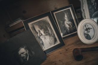 A collection of framed black and white photographs is displayed on a wooden surface. The frames contain portraits: a bearded old man in profile, a young woman gazing sideways, and another young woman playing the violin. The setup has a vintage aesthetic, with a dark, moody atmosphere. A tobacco pipe is also visible, adding to the nostalgic feel.