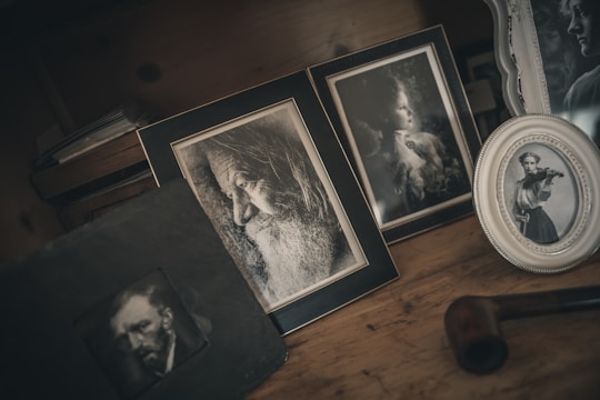 A collection of framed black and white photographs is displayed on a wooden surface. The frames contain portraits: a bearded old man in profile, a young woman gazing sideways, and another young woman playing the violin. The setup has a vintage aesthetic, with a dark, moody atmosphere. A tobacco pipe is also visible, adding to the nostalgic feel.
