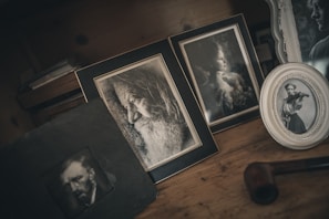 A collection of framed black and white photographs is displayed on a wooden surface. The frames contain portraits: a bearded old man in profile, a young woman gazing sideways, and another young woman playing the violin. The setup has a vintage aesthetic, with a dark, moody atmosphere. A tobacco pipe is also visible, adding to the nostalgic feel.