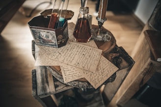 Several handwritten letters are sprawled across a rustic wooden table along with a vintage-looking metallic container that holds glass test tubes filled with colored liquids. A decanter filled with a dark liquid is nearby, next to an ornate candlestick.
