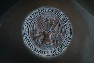 A bronze-colored emblem representing the Department of the Army, United States of America, featuring various military insignia including a helmet, flags, a cannon, cannonballs, and a sword. The emblem includes detailed engravings and the date 1775.