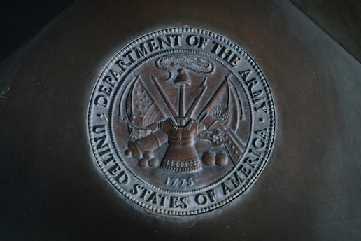 A bronze-colored emblem representing the Department of the Army, United States of America, featuring various military insignia including a helmet, flags, a cannon, cannonballs, and a sword. The emblem includes detailed engravings and the date 1775.