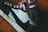 A close-up view of a sneaker featuring a black swoosh design on white leather with dark brown overlays. The shoe has pink laces and is photographed on a wooden floor, highlighting the texture and details of the leather material.