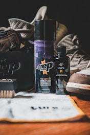 Various shoe care products are arranged on a surface, including two bottles labeled 'crep protect' and 'crep cure,' a brush, a cloth, and a black pouch. The background features a pair of casual sneakers.