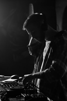 A DJ wearing headphones is focused on mixing music, using turntables and various audio equipment. The scene is captured in black and white, creating a dramatic and engaging atmosphere.