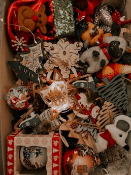 A collection of Christmas decorations is displayed, including plush reindeer, snowmen, and various ornaments. The decorations feature a festive theme with designs such as Christmas trees, stars, and snowflakes made from different materials including wood and fabric. Some of the ornaments are adorned with sequins and glitter, contributing to a festive and sparkling ambiance.