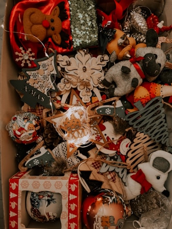 A collection of Christmas decorations is displayed, including plush reindeer, snowmen, and various ornaments. The decorations feature a festive theme with designs such as Christmas trees, stars, and snowflakes made from different materials including wood and fabric. Some of the ornaments are adorned with sequins and glitter, contributing to a festive and sparkling ambiance.