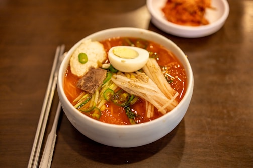 A white bowl filled with spicy Korean cold noodles, known as bibim naengmyeon, topped with sliced cucumber, radish, a boiled egg half, sliced beef, and a dollop of sauce. A pair of metal chopsticks and a spoon rest beside the bowl on a wooden table. In the background, there is a small dish of kimchi.