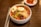 A white bowl filled with spicy Korean cold noodles, known as bibim naengmyeon, topped with sliced cucumber, radish, a boiled egg half, sliced beef, and a dollop of sauce. A pair of metal chopsticks and a spoon rest beside the bowl on a wooden table. In the background, there is a small dish of kimchi.