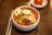 A white bowl filled with spicy Korean cold noodles, known as bibim naengmyeon, topped with sliced cucumber, radish, a boiled egg half, sliced beef, and a dollop of sauce. A pair of metal chopsticks and a spoon rest beside the bowl on a wooden table. In the background, there is a small dish of kimchi.