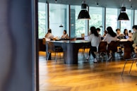 A modern workspace with several people seated around a large, dark table, engaged in work and conversation. The room features large windows, allowing natural light to filter in, and is decorated with overhead pendant lights. The floor has a chevron pattern, adding a warm touch to the sleek interior.
