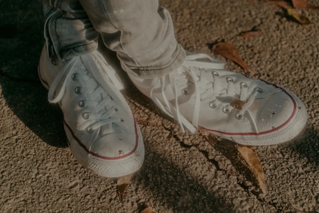 A pair of white canvas sneakers are shown on a rough, textured surface with scattered dried leaves. The laces are loosely tied, and the sneakers have red accents near the rubber soles.