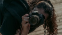 A person with curly hair, wearing a black shirt, is holding a DSLR camera close to their face, appearing to take a photograph. The background is a blurred, wooden texture, suggesting an outdoor setting.