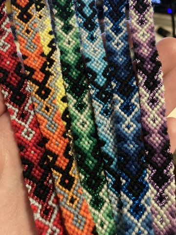 A collection of colorful, intricately woven friendship bracelets featuring geometric patterns. The designs incorporate a variety of colors, arranged in a repeating zigzag pattern, showcased against a person's palm, highlighting the handmade nature of the items.
