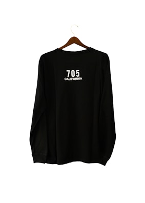 A black long-sleeve shirt is hanging on a wooden hanger. The text '705 CALIFORNIA' is printed in white on the back of the shirt.