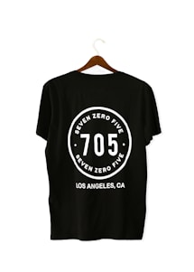 A black T-shirt is displayed on a wooden hanger with a bold white print on the back. The print includes the numbers '705' in large font, encased within a circular design. Above and below the numbers, the words 'SEVEN ZERO FIVE' and 'LOS ANGELES, CA' are printed respectively.
