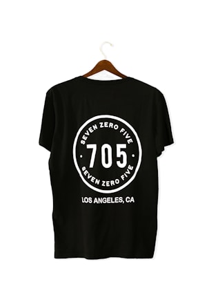 A black T-shirt is displayed on a wooden hanger with a bold white print on the back. The print includes the numbers '705' in large font, encased within a circular design. Above and below the numbers, the words 'SEVEN ZERO FIVE' and 'LOS ANGELES, CA' are printed respectively.