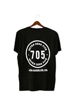 A black T-shirt is displayed on a wooden hanger with a bold white print on the back. The print includes the numbers '705' in large font, encased within a circular design. Above and below the numbers, the words 'SEVEN ZERO FIVE' and 'LOS ANGELES, CA' are printed respectively.