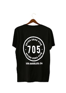 A black T-shirt is displayed on a wooden hanger with a bold white print on the back. The print includes the numbers '705' in large font, encased within a circular design. Above and below the numbers, the words 'SEVEN ZERO FIVE' and 'LOS ANGELES, CA' are printed respectively.