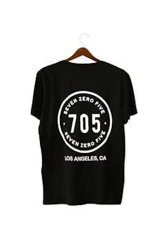 A black T-shirt is displayed on a wooden hanger with a bold white print on the back. The print includes the numbers '705' in large font, encased within a circular design. Above and below the numbers, the words 'SEVEN ZERO FIVE' and 'LOS ANGELES, CA' are printed respectively.