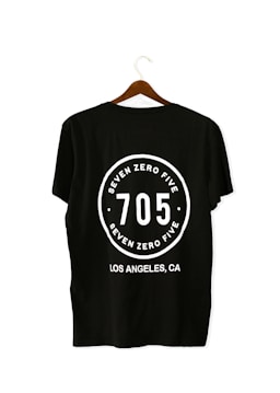A black T-shirt is displayed on a wooden hanger with a bold white print on the back. The print includes the numbers '705' in large font, encased within a circular design. Above and below the numbers, the words 'SEVEN ZERO FIVE' and 'LOS ANGELES, CA' are printed respectively.