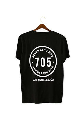 A black T-shirt is displayed on a wooden hanger with a bold white print on the back. The print includes the numbers '705' in large font, encased within a circular design. Above and below the numbers, the words 'SEVEN ZERO FIVE' and 'LOS ANGELES, CA' are printed respectively.