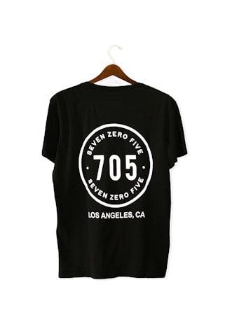 A black T-shirt is displayed on a wooden hanger with a bold white print on the back. The print includes the numbers '705' in large font, encased within a circular design. Above and below the numbers, the words 'SEVEN ZERO FIVE' and 'LOS ANGELES, CA' are printed respectively.