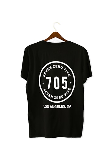 A black T-shirt is displayed on a wooden hanger with a bold white print on the back. The print includes the numbers '705' in large font, encased within a circular design. Above and below the numbers, the words 'SEVEN ZERO FIVE' and 'LOS ANGELES, CA' are printed respectively.