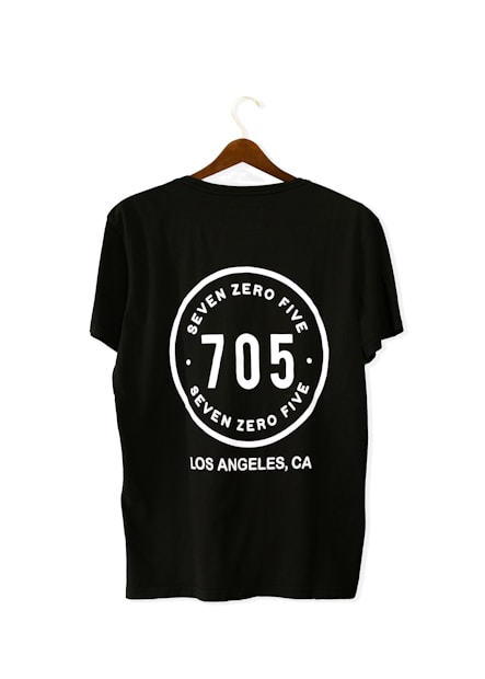 A black T-shirt is displayed on a wooden hanger with a bold white print on the back. The print includes the numbers '705' in large font, encased within a circular design. Above and below the numbers, the words 'SEVEN ZERO FIVE' and 'LOS ANGELES, CA' are printed respectively.