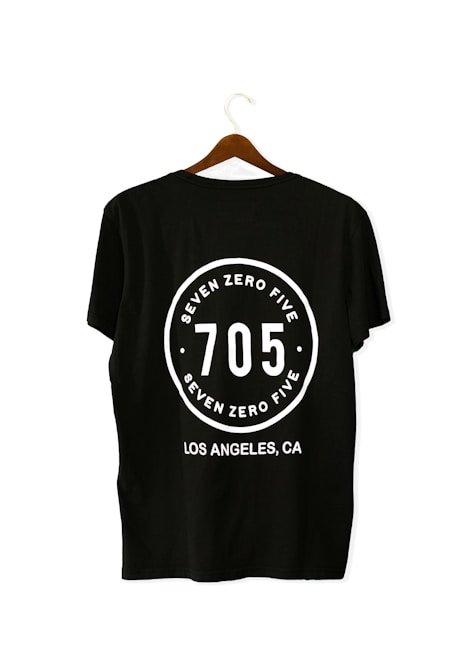 A black T-shirt is displayed on a wooden hanger with a bold white print on the back. The print includes the numbers '705' in large font, encased within a circular design. Above and below the numbers, the words 'SEVEN ZERO FIVE' and 'LOS ANGELES, CA' are printed respectively.
