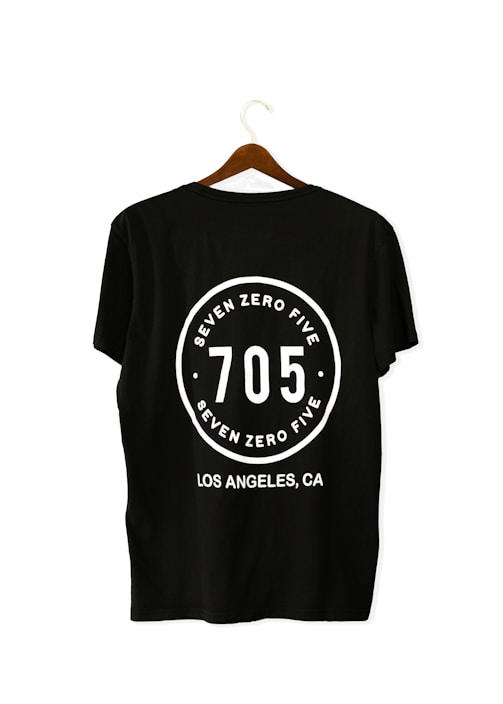 A black T-shirt is displayed on a wooden hanger with a bold white print on the back. The print includes the numbers '705' in large font, encased within a circular design. Above and below the numbers, the words 'SEVEN ZERO FIVE' and 'LOS ANGELES, CA' are printed respectively.