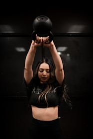 A person is lifting a black kettlebell overhead with both hands. The individual is wearing a dark crop top and appears focused on the exercise. The background is dimly lit, creating a dramatic atmosphere.