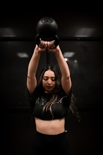 A person is lifting a black kettlebell overhead with both hands. The individual is wearing a dark crop top and appears focused on the exercise. The background is dimly lit, creating a dramatic atmosphere.
