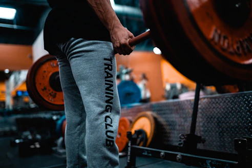 A person wearing grey sweatpants with the words 'TRAINING CLUB' printed on the side is holding a barbell in a gym setting. The barbell is loaded with large, orange weight plates. The background shows gym equipment and has a blurred effect, indicating a focus on the person and the barbell. The environment is industrial, with metallic surfaces and equipment visible.